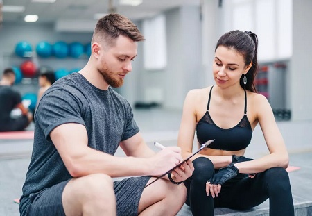 Osoba siedząca na ławce w sali fitness wypełnia formularz na podkładce z dokumentami. Obok siedzi druga osoba w stroju sportowym, obserwująca dokument. W tle widoczne są piłki treningowe i wyposażenie siłowni.