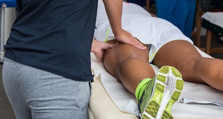 Zabieg manualny wykonywany na nodze osoby leżącej na stole rehabilitacyjnym. Widoczne dłonie terapeuty uciskające okolicę kolana oraz wyprostowana noga w obuwiu sportowym. Zdjęcie przedstawia rehabilitację narządu ruchu w gabinecie fizjoterapeutycznym.